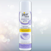 A photo showcases a bottle of Pjur Med Sensitive Glide - Water-Based Lube 100ml (3.4oz) by Pjur. The product features purple and white packaging that highlights its water-based formulation, making it perfect for addressing vaginal dryness and protecting sensitive mucous membranes. It is free from glycerin, parabens, and preservatives and is manufactured in Germany.